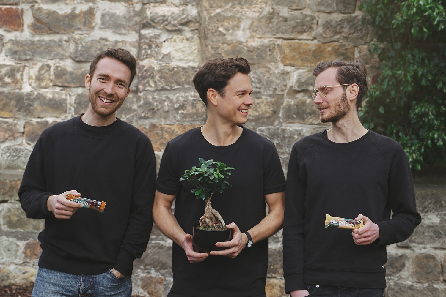three young men in front of a wall with granula bars in hand and a plant that looks like a tree